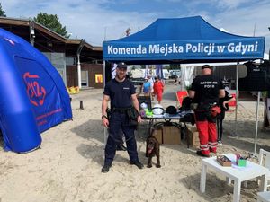 Zdjęcie przedstawiające policjanta z psem służbowym oraz inne osoby podczas festynu na plaży miejskiej.