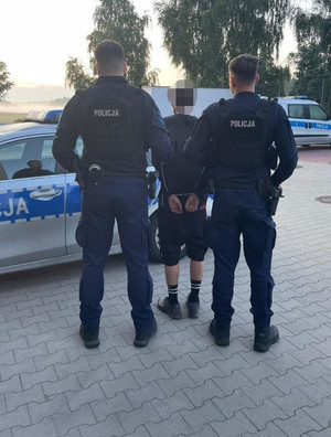 Zdjęcie przedstawiające dwóch policjantów z zatrzymanym mężczyzną na tle radiowozów policyjnych.