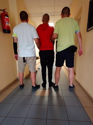 Zdjęcie przedstawiające dwóch policjantów prowadzących zatrzymanego mężczyznę.