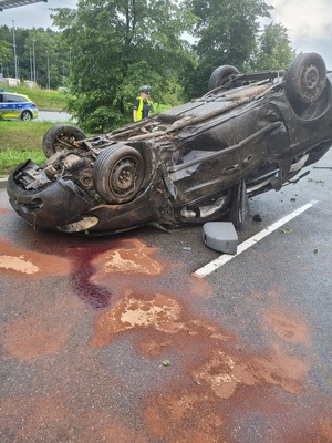 Zdjęcie przedstawiające pojazd biorący udział w wypadku drogowym. Pojazd jest usytuowany na dachu, na ulicy. Na zdjęciu widać policjantkę i część radiowozu policyjnego.