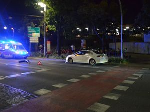 Zdjęcie przedstawiające hulajnogę, leżącą na ulicy, biały pojazd osobowy oraz karetkę pogotowia.