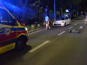 Zdjęcie przedstawiające hulajnogę, leżącą na ulicy,  biały pojazd osobowy, karetkę pogotowia oraz mężczyznę idącego przed pojazdem.