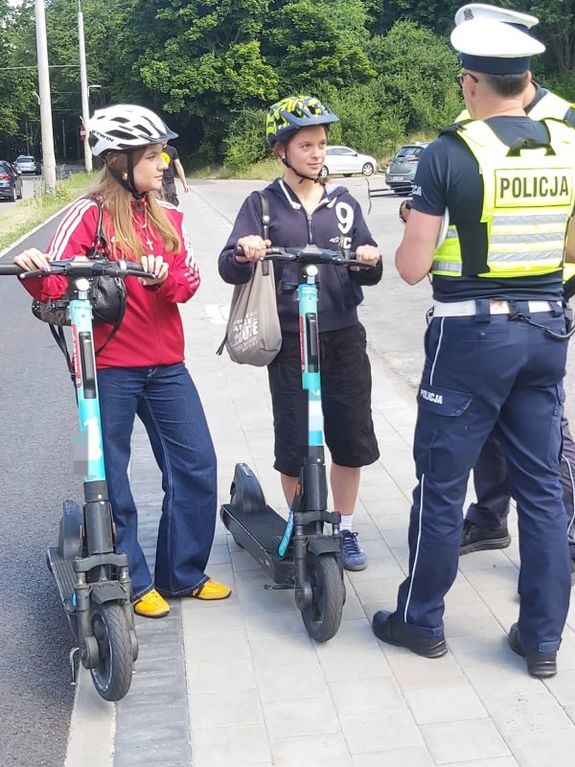 Zdjęcie przedstawiające dwóch policjantów podczas rozmowy z dwiema nastolatkami, które korzystają z hulajnóg.