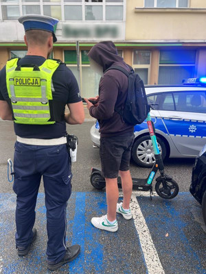 Zdjęcie poglądowe, przedstawiające na tle radiowozu policjanta z użytkownikiem hulajnogi.