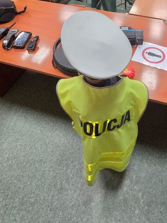 Zdjęcie przedstawiające przedszkolaka stojącego tyłem do aparatu. Dziecko ma na sobie kamizelkę odblaskową i policyjną czapkę.